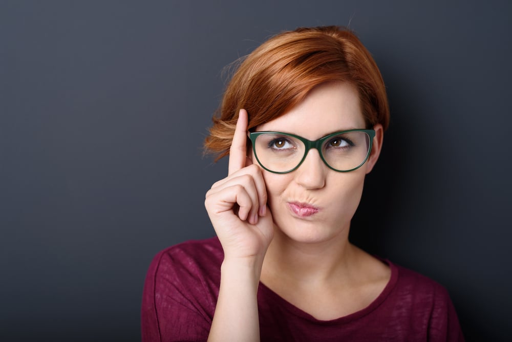 Nerdy scholastic young woman wearing geeky glasses standing thinking with her finger raised and a grimace of concentration in a humorous stereotypical depiction, over a dark background with copyspace